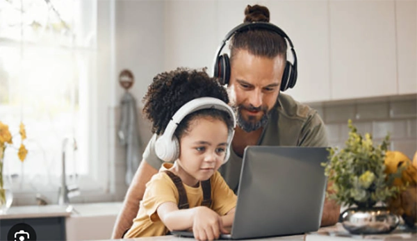 A child wearing headphones at a laptop, generating a song on an AI music platform while a parent looks on.